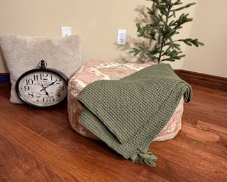 A handwoven pouffe featuring a geometric design in soft pink, cream, and light gray shades. The pouffe has a square shape with decorative patterns along the edges. The fabric is textured, and it appears to be made of natural materials.

Also included in this decorative collection is a green waffle textured throw, a clock that can be converted for either mantle or wall use and a decorative tree. 