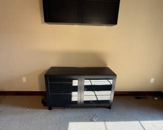 This item features a sleek black finish with a contemporary design. It includes a TV stand with glass sliding doors and multiple shelves for storage. The stand is made from a pressed wood material with a sturdy structure. The top section is a matching black cabinet with two compartments, designed for wall mounting. There are visible logos and labels indicating the brand, along with metal hardware for assembly