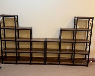 The item features two multi-level shelving unit with a modern industrial design. It has a combination of a dark metal frame and wood finish shelves with a rich brown tone. The shelves are open and rectangular in shape, displaying an X-brace design on the sides. The unit consists of three taller sections on either side and connecting lower shelves, creating a staggered effect. Measuring tape visible on the shelves indicates dimensions for potential storage use