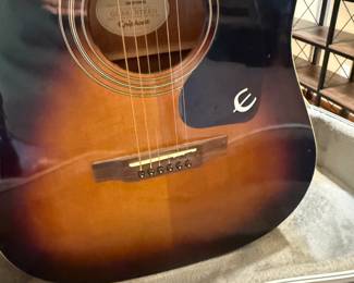 Black acoustic guitar with a glossy finish featuring a sunburst design. It includes colorful decorative stickers of a shield and a deer, positioned on the body. The brand name 'Ibanez' is displayed on the headstock and the label inside the guitar indicates the model AEG20II-TRS-BR-01. The fretboard is made of dark wood with gold tuners, and the guitar is housed in a fitted hard case with a soft interior lining.