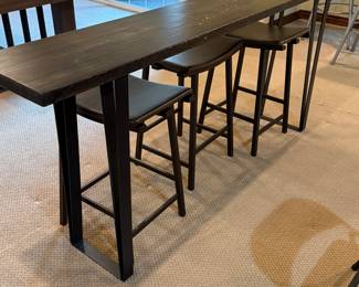 This set features a long, rectangular bar height table with a smooth dark wood surface. The table legs are made of sturdy black metal, providing a modern contrast. Accompanying the table are three stools with a sleek black frame and rich brown padded seats, designed for comfort. The stools have a curved seat shape for added ergonomics. Three Bar Stools. 