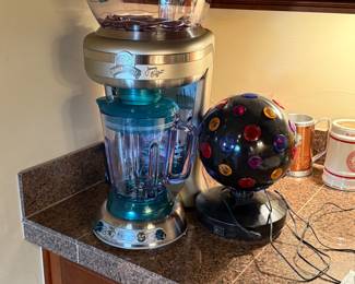 The item features a retro style glass blender with a teal base and a transparent blending jar. The controls are metallic silver with teal accents. There is an ice processor attachment on top, which is purple and clear. Also included is a decorative disco ball, black in color with multi-colored circular decorations, mounted on a black base.