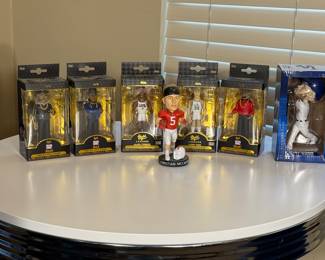 A group of eight premium vinyl figures showcased in packaging. Includes figures of Run DMC, Allen Iverson wearing a white jersey, Larry Bird in a Celtics uniform, Christian McCaffrey in a red jersey with the number 5, and a bobblehead of a player in a white uniform with a blue cap. The figures feature various colors such as black, blue, white, and red, and display brand logos. Each figure is encased in transparent packaging with branding details visible.