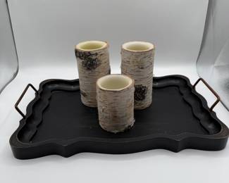 Three cylindrical candle holders resembling birch logs with a creamy interior. The candle holders have a natural bark texture and a light color with darker patches. They are placed on a decorative black tray with a wavy edge and two metal handles.