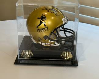 A gold mini helmet with a black stripe, featuring the Heisman Trophy logo on one side and autographed by Tony Dorsett. The helmet has an autograph by a Heisman winner on the top, along with the inscription 'Heisman 76'. The helmet is displayed in a clear acrylic case on a black pedestal with gold accents, which includes an engraved plate that reads 'HEISMAN TROPHY FRESHMAN OF THE YEAR TONY DORSETT.”