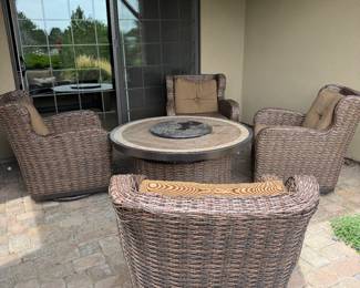 Set includes four woven wicker chairs with brown cushioned seats and a circular fire pit table featuring a stone top. The chairs have a rounded shape, and the cushions are a solid brown color with button detailing. The fire pit table has a dark metal rim and a circular opening filled with decorative glass stones.