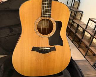 An acoustic guitar with a natural wood finish, featuring a black pickguard and six strings. The guitar is identifiable by the brand name 'Taylor' on the headstock. Accompanied by two guitar cases: a vintage brown hardshell case and a the original tan soft case, each with zippers and straps.