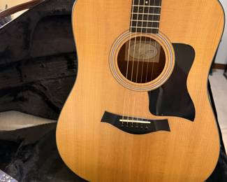 An acoustic guitar with a natural wood finish, featuring a black pickguard and six strings. The guitar is identifiable by the brand name 'Taylor' on the headstock. Accompanied by two guitar cases: a vintage brown hardshell case and a the original tan soft case, each with zippers and straps.