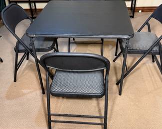 A square black folding table accompanied by four black folding chairs. Chairs feature gray padded seats with a mesh pattern. All items are made of metal, with a sleek, modern design.
