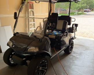 
EVolution Black electric golf cart with a canopy top, featuring two-tone seating in black and gray. The cart has custom black wheels with a stylish design, a steering wheel with a silver center logo, and various dashboard controls. The exterior includes a clear windshield and headlights, with visible wiring inside. Powers on and appears to be in good working order. See photos for front end damage. 