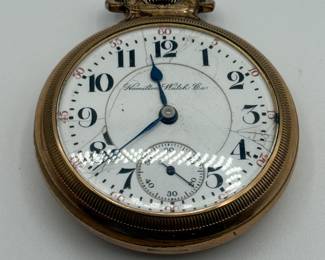A vintage pocket watch displayed under a clear glass dome. The watch has a round silver case with an intricate design on its edge. The dial features black Arabic numerals and blue hour and minute hands, with a small subsidiary seconds dial at the bottom. The brand name 'Hamilton Watch Co.' is elegantly printed near the top of the dial. The wooden base supporting the dome is light brown in color.