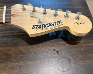 A black electric guitar with a glossy finish and a white pickguard. The body is shaped in a classic style. The headstock is natural wood with the text 'Starcaster' and 'Fender' branded logo. The fretboard is dark brown with metal fret markers. Accompanying the guitar is a black gig bag with the logo 'Epiphone' displayed on it.
