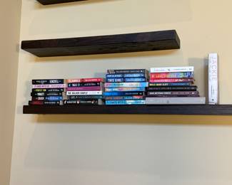 A collection of books displayed on a dark wood floating shelf. The books vary in colors, including black, pink, white, and blue. Some titles visible include 'It Ends With Us' by Colleen Hoover, 'The Inmate' by Frieda McFadden, and 'Ugly Love' by Colleen Hoover. The shelf shows a mix of genres and authors, featuring both hardcover and paperback editions. A small book titled 'Jackie' is also present on the shelf, standing out with its white cover.