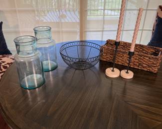 Three clear glass jars with a slight blue tint, a round black metal wire bowl, a rectangular woven basket made of natural fibers, and two rustic candle holders featuring twisted candles in a soft pink color, with round wooden bases.