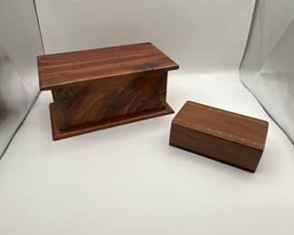 This set includes two wooden boxes; one larger with a rectangular shape featuring a rich, dark wooden grain and a smooth finish, highlighted by light and dark streaks. The corners are detailed with metal brackets. The lid opens to reveal a spacious interior. The smaller box is also rectangular with a lighter wood tone, textured with fine lines along the top and a clean, polished finish