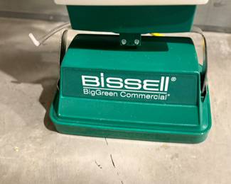 This floor care machine features a green base with a white upper section. It is branded with 'Bissell' and 'BigGreen Commercial'. The machine includes a long handle and comes with multiple brush accessories in white, brown, and yellow colors. Additionally, a metal plate on the machine displays the model 'P-2100' and series number '72-131K'.

