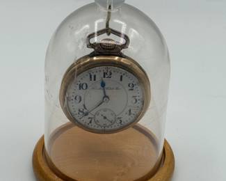 A vintage pocket watch displayed under a clear glass dome. The watch has a round silver case with an intricate design on its edge. The dial features black Arabic numerals and blue hour and minute hands, with a small subsidiary seconds dial at the bottom. The brand name 'Hamilton Watch Co.' is elegantly printed near the top of the dial. The wooden base supporting the dome is light brown in color.