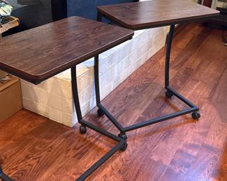 Two side tables featuring a dark wood finish top and sturdy black metal frames. Each table has a rectangular shape with a smooth surface and measures approximately 24 inches wide. The tables are supported by rolling wheels for mobility.