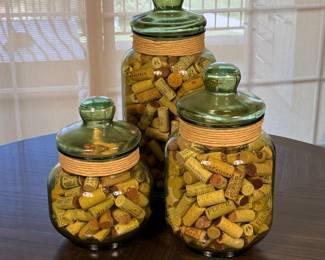 Three vintage glass jars in green glass with clear lids. They are filled with assorted wine corks in various colors, including yellow, brown, and natural cork tones. Each jar features a textured, ribbed design on the body and is secured with a light tan rope band around the neck.