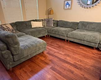 A large sectional sofa in a soft green corduroy fabric, featuring plush cushions. The sofa has a sectional shape with a chaise lounge on one side. Wooden legs in a natural finish. There are decorative throw pillows in a mix of colors and patterns, including a plaid design. Each section can be separated and each section is approximately 45" wide. 