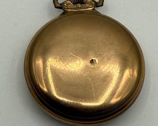 A vintage pocket watch displayed under a clear glass dome. The watch has a round silver case with an intricate design on its edge. The dial features black Arabic numerals and blue hour and minute hands, with a small subsidiary seconds dial at the bottom. The brand name 'Hamilton Watch Co.' is elegantly printed near the top of the dial. The wooden base supporting the dome is light brown in color.