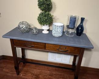Console table with a gray surface and wooden legs. Features two drawers with metal handles. The tabletop is adorned with two decorative geometric spheres in silver, a white planter with a double ball topiary, a decorative blue and white patterned container, a clear glass candle holder with a dark blue candle, and a dark blue vase.