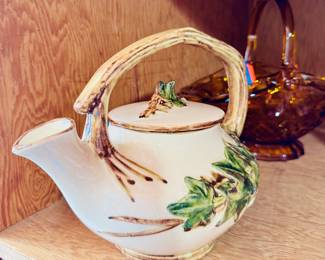 Cream-colored ceramic teapot with an arched handle designed to look like a tree branch. Green leaves and brown bark details are hand-painted and slightly raised. Lid has a matching branch and leaf motif.
	•	Style: Mid-century rustic