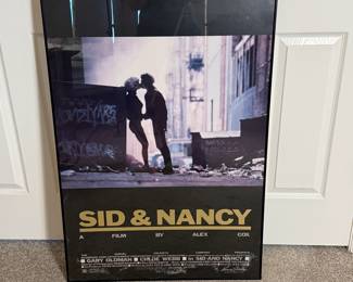 The item features a framed movie poster for 'Sid & Nancy,' showcasing a silhouette of a couple against a backdrop of urban graffiti. The poster includes text in large, bold yellow letters at the bottom, stating 'SID & NANCY' along with the words 'A FILM BY ALEX COX.' It displays the names 'GARY OLDMAN' and 'CHLOE WEBB' prominently. The frame is black, and the overall dimensions appear to be significant, as indicated by the measuring tape in the images.