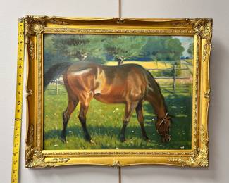 A framed signed painting, naive, of a glossy bay horse wearing a halter and grazing in a grassy field, with wooden fences, roads and orchard in background
