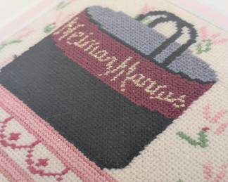 Closeup of the charming framed needlepoint scene of a shopper's closet, showing coats, dresses, purses and hats, with shopping bags from Neiman Marcus and Saks.