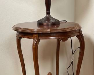 Glass shade lamp depicting a scene reminiscent of a Thomas Kinkade painting. Heavy bronze base. Powers on. H 22" x 13" diameter. Wood accent table, no markings. H 24" x 19" diameter. Small crack under the top.