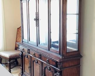 Ashley Classical designed wood veneer dining hutch featuring carved edges, (2) levels of glass shelving, (3) drawers and (2) lower cabinet doors. H 78" x W 70" x D 18". Matches lot 101. Objects shown in background are not included. Receipt from consignor shows Ashley as the brand of furniture.