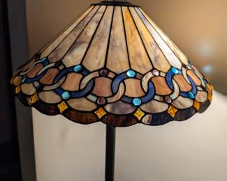 A stunning and elegant floor lamp in the classic Tiffany style. The lampshade features a beautiful, multi-colored stained glass design with intricate patterns and cabochon glass jewels. This lamp provides a warm, inviting glow and would be a beautiful, artistic focal point for any living room, library, or den.