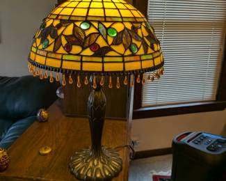 A stunning and elegant lamp in the classic Tiffany style. The lampshade features an intricate stained glass design of leaves and berries, accented with beautiful hanging glass jewels. The warm, inviting glow from the glass makes this a perfect artistic focal point for any room. This is a quality reproduction lamp, ready to be the highlight of your home decor.
