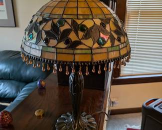 A stunning and elegant lamp in the classic Tiffany style. The lampshade features an intricate stained glass design of leaves and berries, accented with beautiful hanging glass jewels. The warm, inviting glow from the glass makes this a perfect artistic focal point for any room. This is a quality reproduction lamp, ready to be the highlight of your home decor.