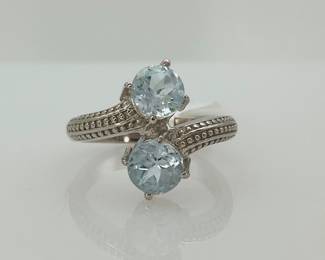Two (2) round cut 5.7 mm light blue topaz stones, 1.70 ctw, sit on either side of a sterling silver rope design ring. Size 7. 4.92 gtw. Includes certificate provided by consigner.