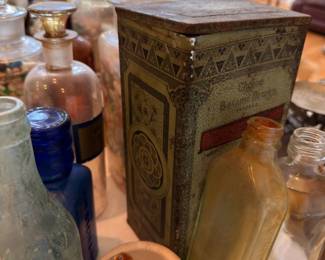 Ornate embossed metal tin with hinged lid and decorative pattern; surrounded by antique glass bottles including aqua and clear apothecary jars; unique pharmacy and general store collectible.