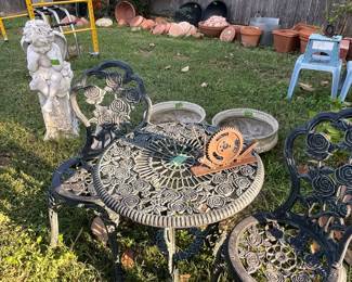 Very nice, heavy metal three-piece yard table and two chairs. Meditating angel sitting on pedestal. Newer yellow folding scaffold and Rubbermaid two door storage in background. Lots of pottery against fence.