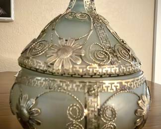 A covered dish made of frosted glass, adorned with silver-colored metalwork in floral and scroll patterns, and a decorative finial on the lid