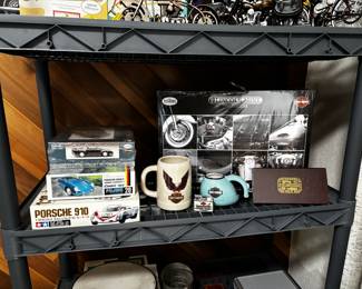 This shelf holds a mix of collectible model kits and Harley-Davidson memorabilia. Items include Porsche model kits (Porsche 356, 910, and Carrera), Harley mugs and keepsakes, a Harley-Davidson Lincoln Mint set (still sealed), and a vintage-style Auto Rallye collectible box. Great mix for auto and motorcycle enthusiasts.