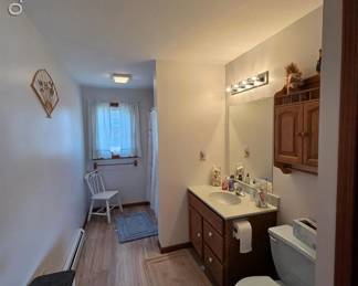 Another bathroom, starting on the left, the hamper, and garbage can, heater, chair and floor rug, vanity and mirror, and light, and toilet paper, and the toilet, along with the medicine cabinet. Floor rugs too