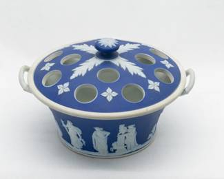 A Wedgwood blue jasperware lidded potpourri pot featuring applied white sprigs creating a bas-relief frieze of classical figures around the outside, and two white handles. The lid also features white bas-relief decoration and a raised finial on the top. A post-1930s mark to base of “WEDGWOOD” and “Made in England” and potter’s mark. 4.5” tall to height of knob, 7” across handle to handle, 6” across the rim. 