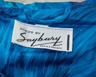 Closeup of a darling vintage A-line belted dress in swirling rich blues and violet pattern, rhinestone button front and with pockets; label reads Styled by Saybury 