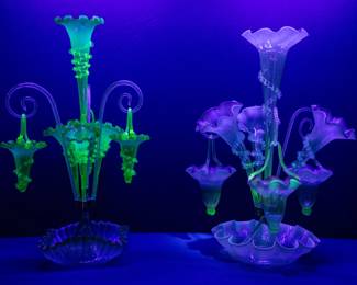Seen under black light: Antique Victorian era art glass epergnes or centerpiece vases, the first a 4-trumpet epergne with 3 hanging baskets, cranberry uranium vaseline glass; trumpet horns with ruffled top rim and hand applied clear rigaree on the neck, the baskets with ruffled rims and hand applied handles above a round base with crimped edges. The second antique 19th century art glass epergne has three vaseline uranium glass trumpets around a taller central trumpet, and three spiral canes suspending three baskets, all with crimped ruffled edges above a squared base with crimped edges.