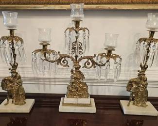 Antique French Brass & Crystal Girandole Mantel Candelabra Set with Marble Bases. Set of three ornate Louis XVI–style girandole mantel candelabras, crafted in brass and adorned with hanging cut glass prisms. Each rests on a white marble base with felt bottoms and features figural scenes in classical dress. The tallest measures approximately 18–20" high. Early 20th century in style and execution, this three-piece set is both decorative and functional, with glass hurricane shades adding extra elegance.