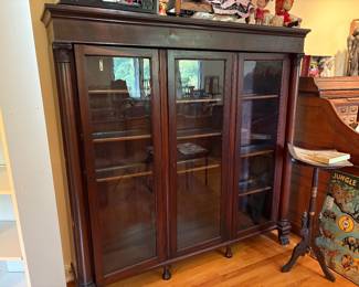 American Empire Revival Bookcase/Display Cabinet, dating to the mid-to-late 19th century. Crafted from rich, dark wood, likely mahogany, three tall glass doors that lock with a key, flanked by impressive carved columns with detailed capitals. The cabinet is supported by intricately carved front paw feet, a hallmark of the Empire style.