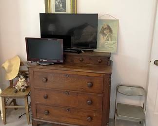 A folding chair and a wooden chair sit next to this three-drawer dresser and two video monitors.