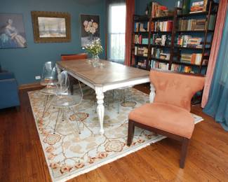Another view of the dining table with two upholstered accent chairs and a set of six mid-century inspired transparent acrylic and chrome  chairs, wooden open frame bookcases, antique and vintage books, a large light color floral theme area rug, framed paintings and prints