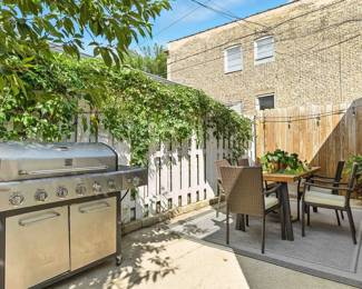 A Kenmore cabinet style 6-burner gas grill with side burner, wooden dining table with set of rattan wicker dining chairs, a large outdoor area rug, and vibrant potted plants with planters