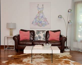 A rich deep brown leather three seat rolled arm sofa, a mid century modern chrome three-arm floor lamp, a mid century modern style high gloss coffee table with glass legs, a cowhide area rug